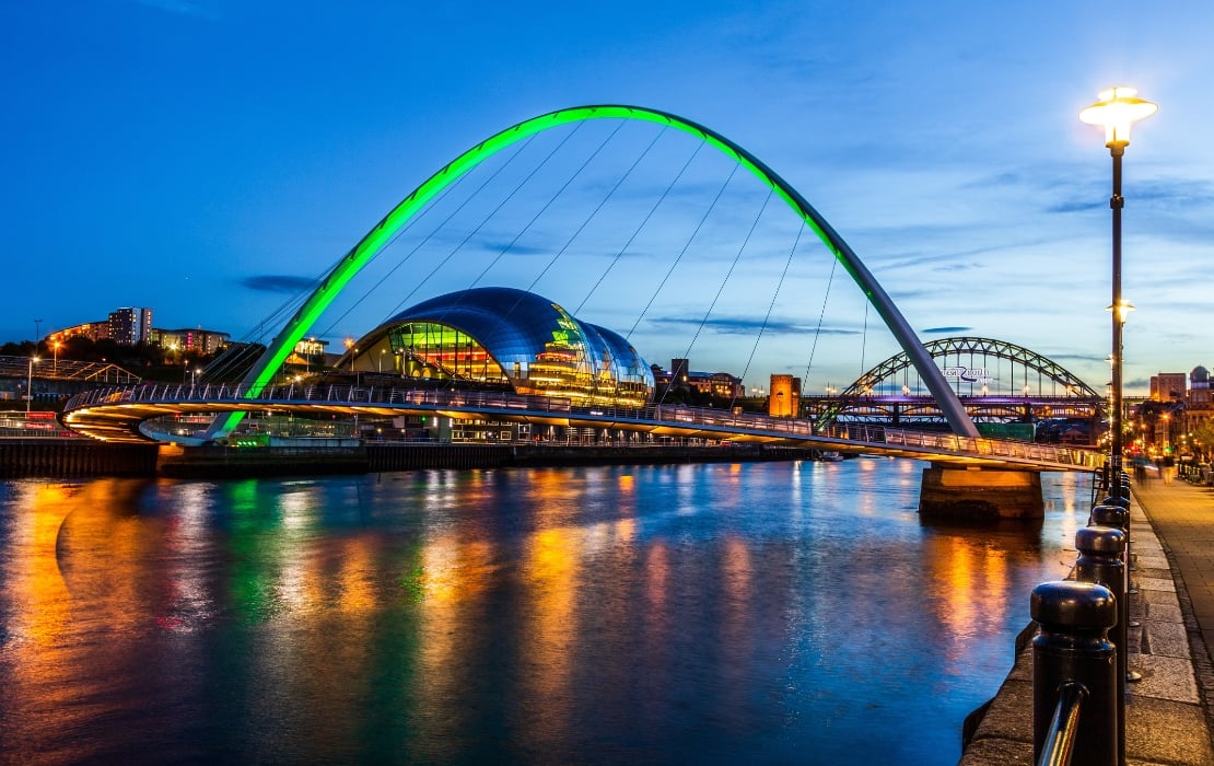 A photo of the Newcastle and Gateshead Millennium bridges over the river Tyne in Newcastle, UK. The photo is taken at sundown, and the Gateshead Bridge is lit up in bright green, with its reflection shining on the water.