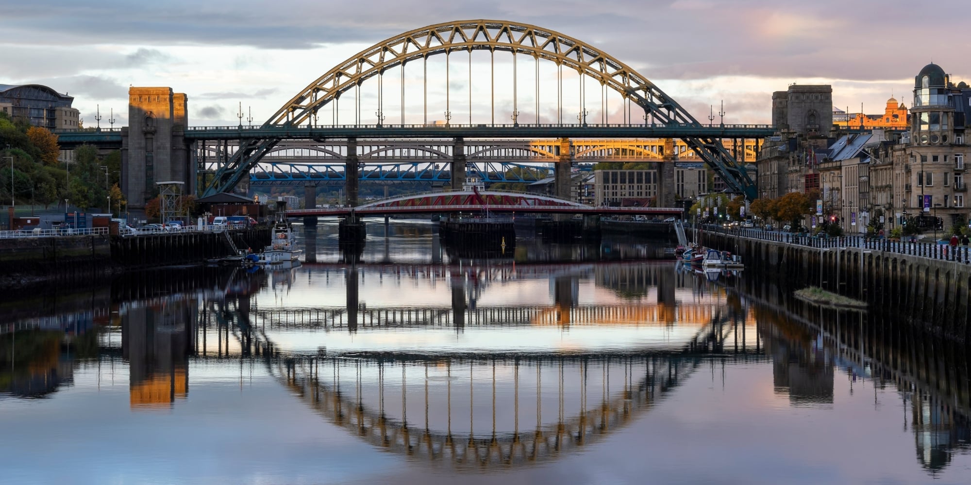 A scenic view of the Tyne Bridge in Newcastle upon Tyne, spanning the River Tyne. The arched steel bridge is reflected in the calm water below, along with two other bridges in the background.