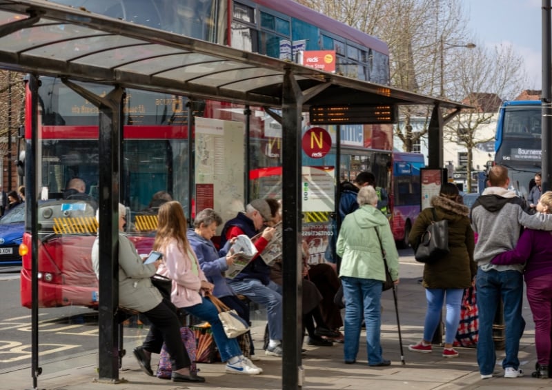 A group of people waiting at a covered bus stop in a busy urban street. Some are seated while others stand, looking toward the road or checking phones.