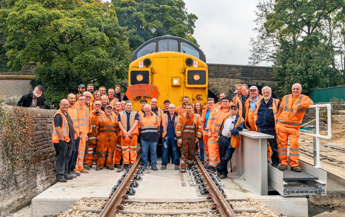 A photo of a large group of volunteers wearing high vis clothing and standing in front of an orange train carriage, on the tracks of the railway bridge they'd been upgrading. There's a few trees behind them.