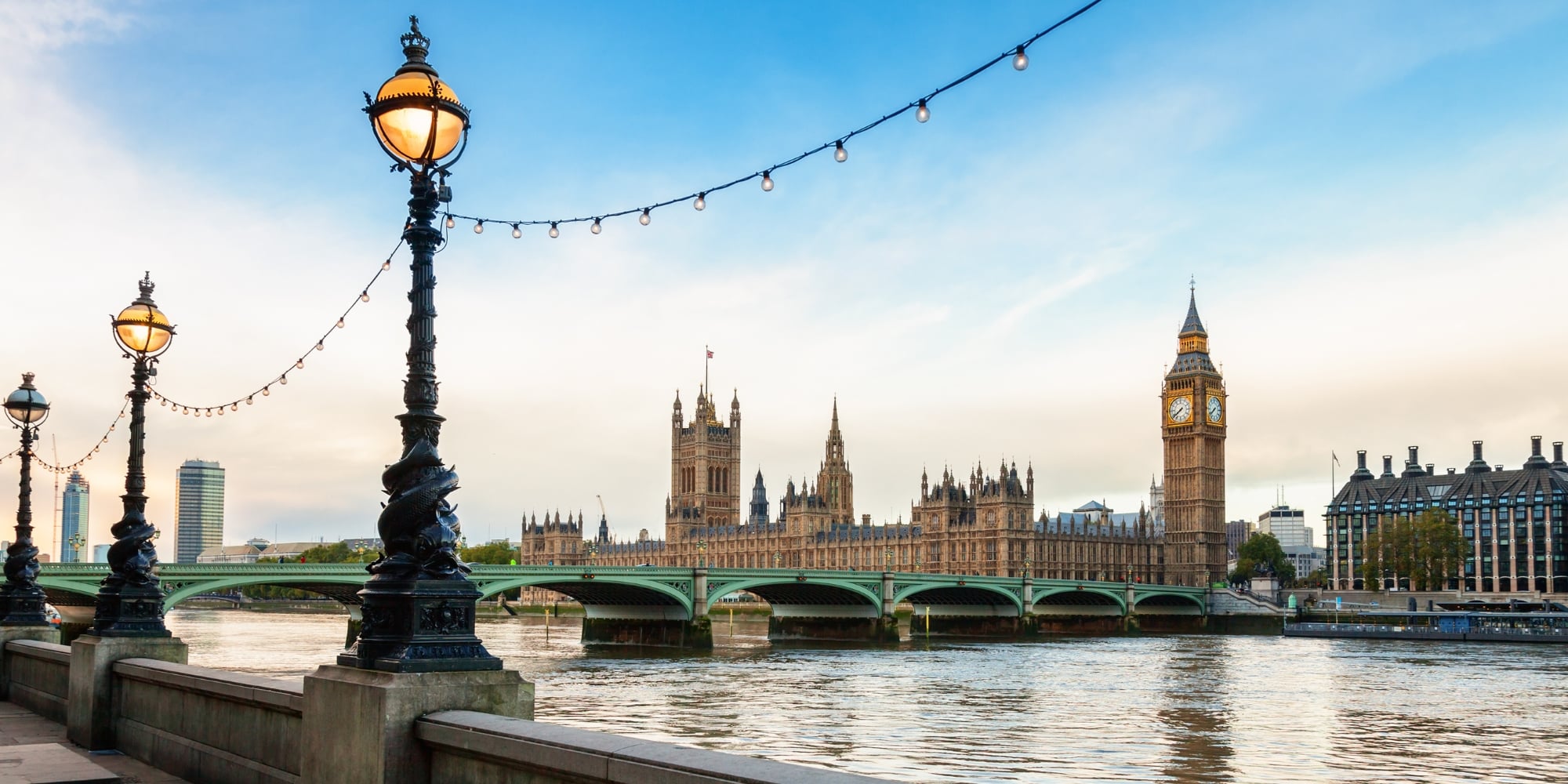 A photo of a London cityscape featuring Westminster Abbey and Big Ben in the background, with the River Thames and lamp posts a dolphin design in the foreground.