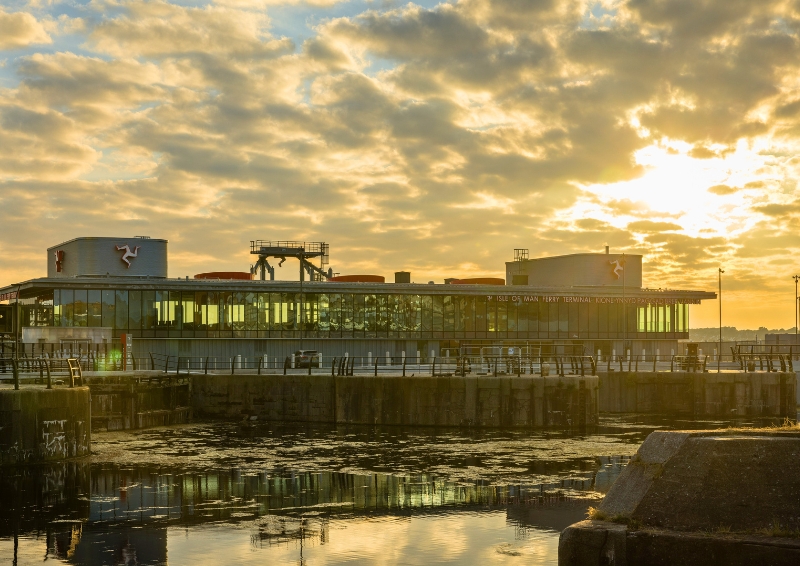 Modern glass-fronted ferry terminal building on a waterfront at sunset, with golden sunlight reflecting off calm water in the foreground and dramatic clouds overhead.