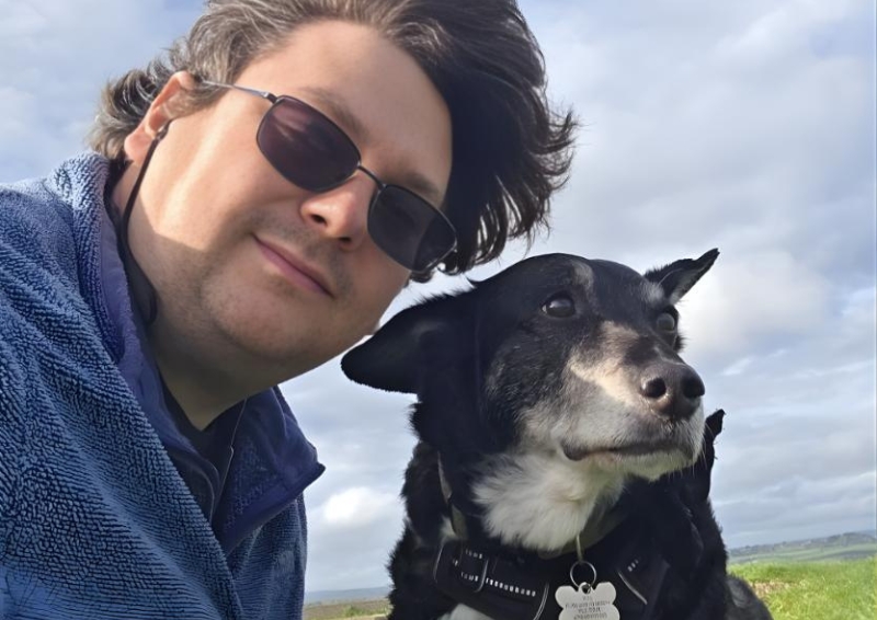 Richard Lang wearing sunglasses and a fleece jacket sits outdoors beside a black dog wearing a collar, with open countryside and a cloudy sky in the background.