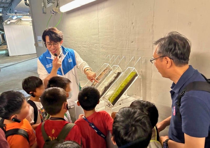 Educator using a headset microphone demonstrates angled tray displays of layered soil or materials to a small group of school children, who are gathered closely around while another adult observes in an indoor workshop setting.