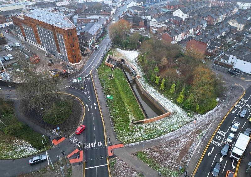 Aerial view of an urban flood management scheme with a narrow engineered water channel running through a landscaped green corridor, bordered by paths and trees, alongside a curved road junction, residential housing, and a modern multi-storey building, with light snow dusting the ground.