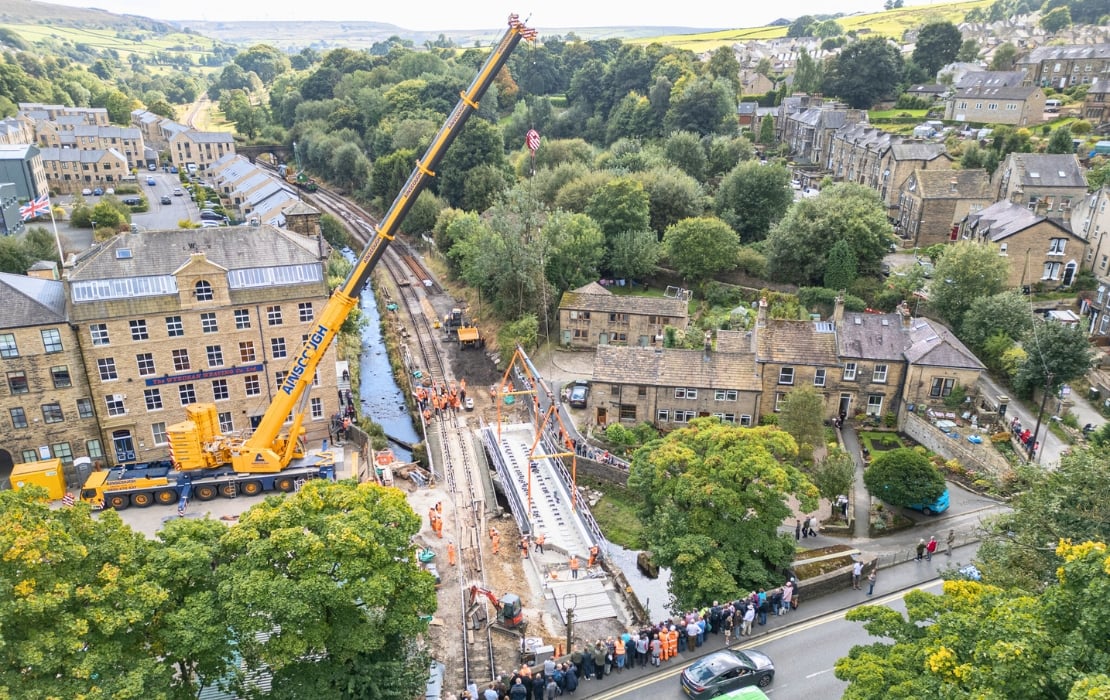 An aerial view photo of a large yellow crane onsite at the Bridge 27 Renewal project. The crane is to the left of the track and is surrounded by a building on the top, and some trees on the bottom. To the right of the tracks, there's more buildings surrounded by trees.