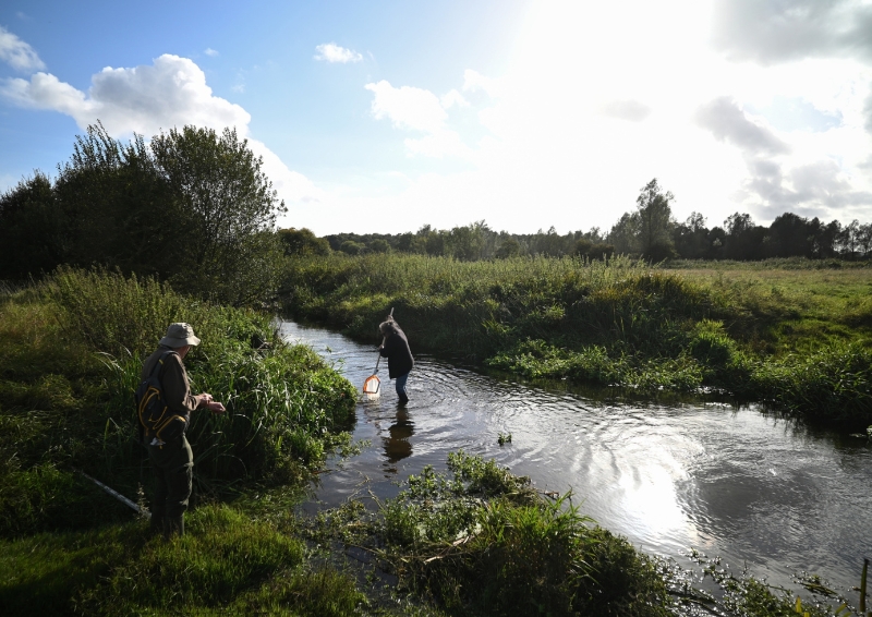 Two people wearing jackets and wellies stand by a river, with nets and other equipment to test the water for pollution. The river is surrounded by fields, grass, shrubs and trees and the sky is bright with some clouds.
