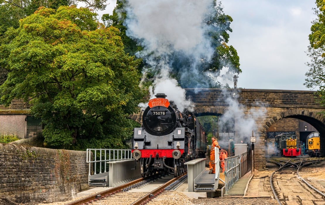 A photo of an old-fashioned steam train with the words 'Bridge 27 Renewal' printed on its front arriving at a station, a cloud of steam emerging from it. There are trees to its left and a bridge to the right. 