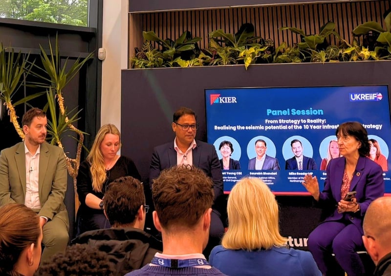 Four panellists sit at the front of a room during a UKREiiF panel session, with an audience seated in the foreground. A large screen behind them displays “Panel Session: From Strategy to Reality” alongside speaker headshots and logos for Kier and UKREiiF.