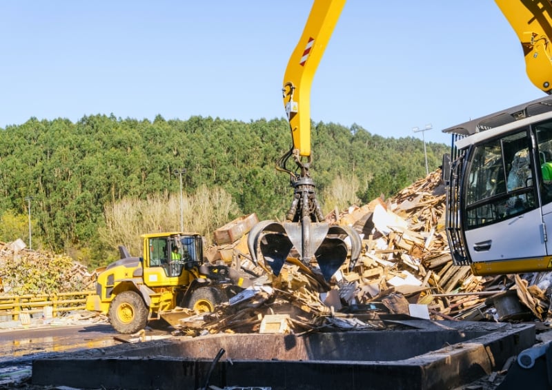 A mechanical claw on an excavator lifts a pile of broken wooden waste at a recycling facility, with construction machinery and a wooded hillside in the background.