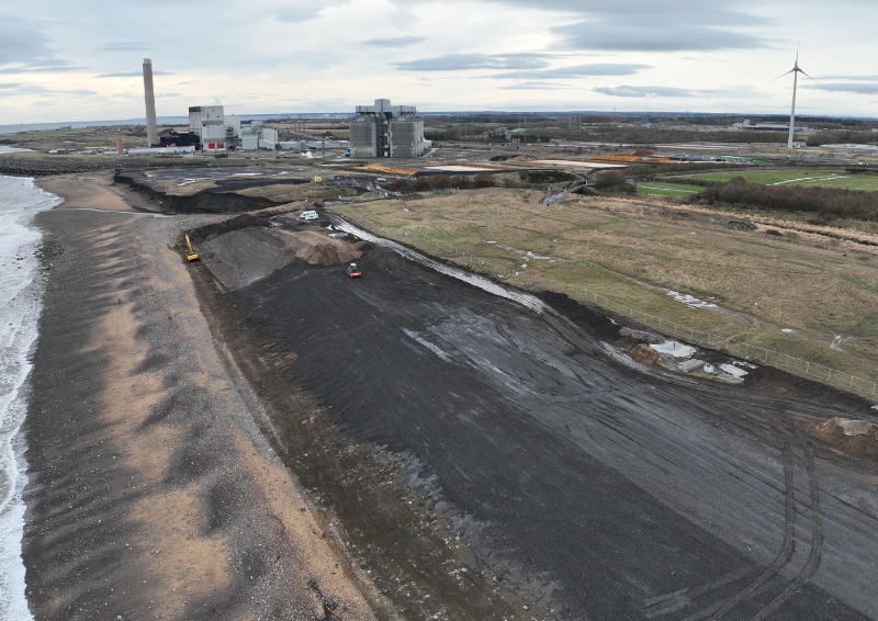 A photo of an aerial view of the Lynemouth Coastal Landfill Remediation project, with a view of the beach, and a few buildings and wind turbine in the background. 