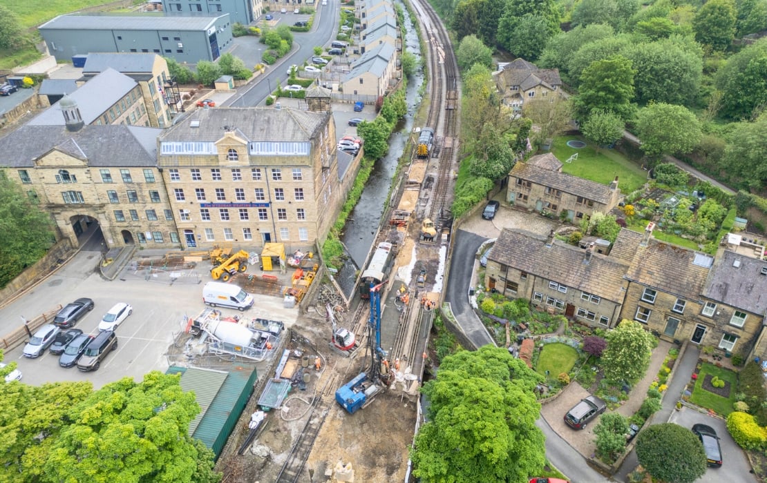 An aerial photograph of the Bridge 27 Renewal project. The tracks of the railway bridge are at the centre of the photo. To the left, a couple of buildings and a car park. To the right, a few smaller buildings surrounded by trees.