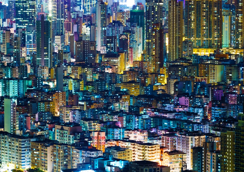 A photo of high rise residential buildings in Hong Kong at night. The buildings are very close together with lights turned on within several of the many windows visible.
