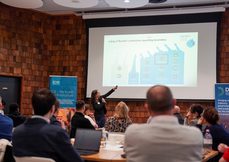 Conference setting with a presenter pointing to a projected slide showing emissions reporting data, while attendees sit at tables facing the screen in a wood-panelled room, with banners and laptops visible.