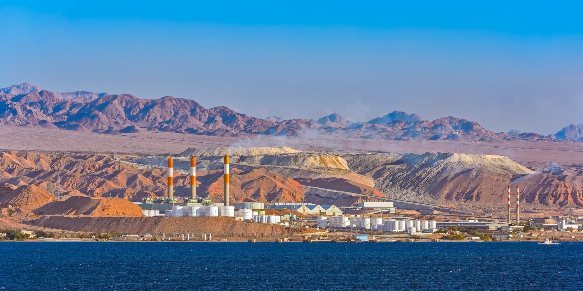 Industrial oil refinery on the Red Sea coast near desert mountains in the Middle East, featuring storage tanks, chimneys, and rugged terrain under a clear blue sky.
