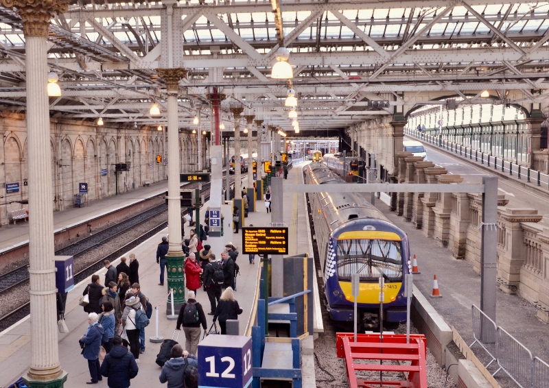 Passengers standing on a covered railway platform as a blue and yellow train waits alongside, with overhead steel beams, platform signs, and departure boards visible inside a large station.