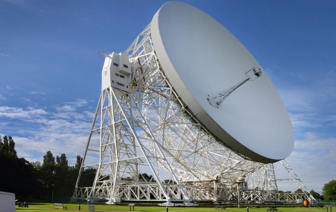 A large white radio telescope dish standing on a complex metal framework in an open grassy area, set against a bright blue sky with scattered clouds.