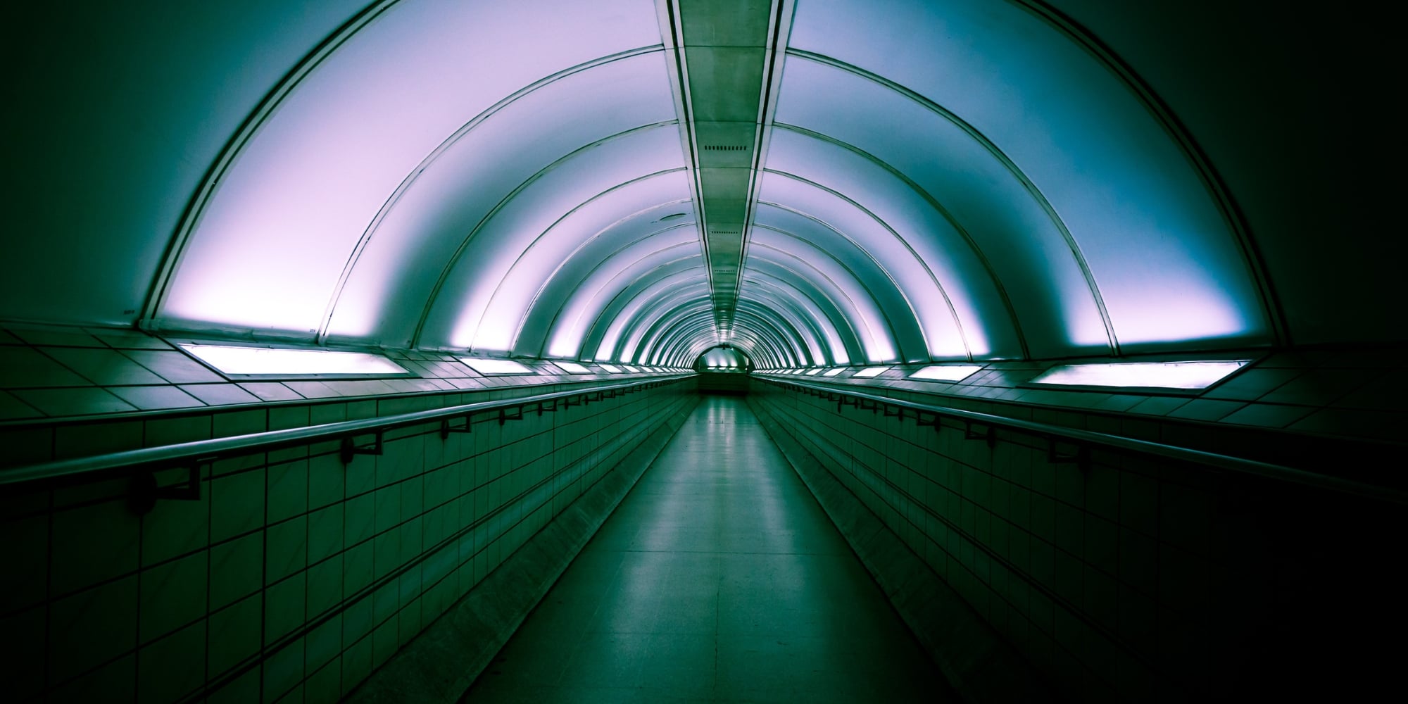 A photograph of a tunnel in Bank Station in London. The bottom half of the wall is tiled, with hand railing about halfway up the image. The top half is lit up by light panels that cast a slightly purple light on the top of the tunnel.