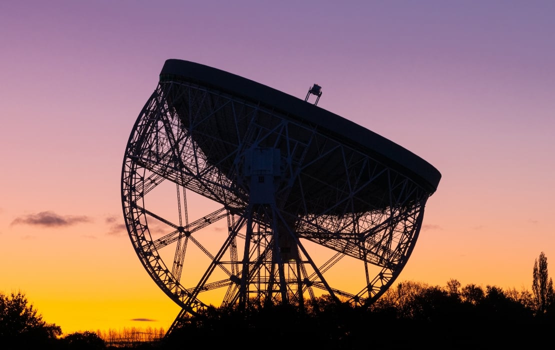 A large radio telescope dish silhouetted against a vibrant sunset sky, with shades of orange, yellow, and purple. The telescope’s metal framework is visible, rising above dark tree silhouettes at the horizon