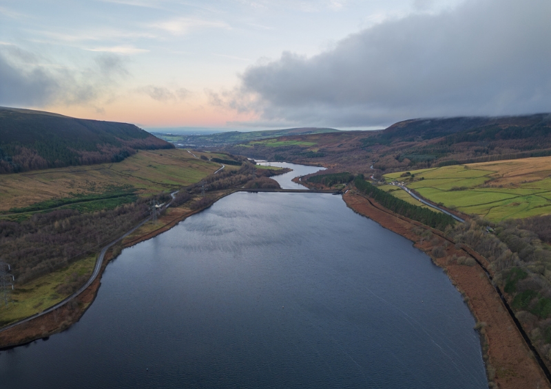 Aerial view of a large reservoir surrounded by rolling hills and green fields. The water stretches into the distance, bordered by winding roads and patches of woodland. 