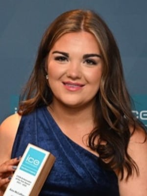 Head-and-shoulders portrait of a person wearing a blue sleeveless outfit and holding an ICE award against a branded event backdrop.