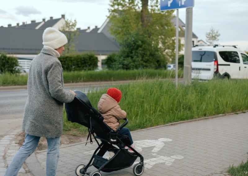 Person pushing a stroller along a paved pedestrian path beside a road, with a child seated inside the stroller, grassy verge and residential houses in the background, and a pedestrian path marking visible on the pavement.