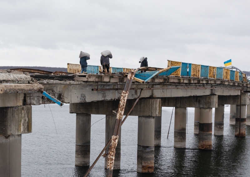 A photo of a few pedestrians carrying filled sacs as they cross a destroyed bridge over Siverskyi Donets in Stary Saltiv, Ukraine. The railings on the bridge are painted in yellow and blue and a Ukrainian flag flies in the background. It's a grey, cloudy day.