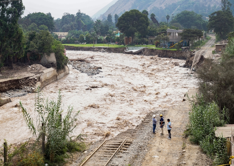 A birds-eye view photograph of an overflowing river caused by heavy rains from the El Niño phenomenon. A few people stand by the flooded river.