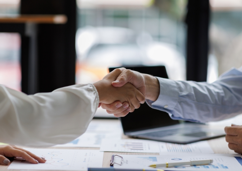 Two people shaking hands across a table in a business setting, with documents, a pen, and an open laptop visible in the foreground.