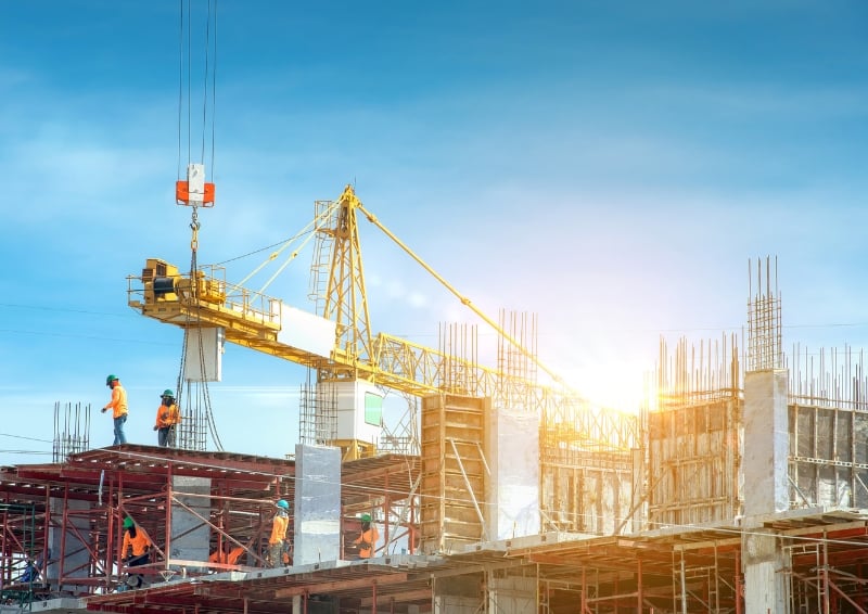 Construction site with workers wearing safety gear on scaffolding, a large yellow tower crane lifting materials, and partially built concrete structures under a clear blue sky with sunlight in the background.