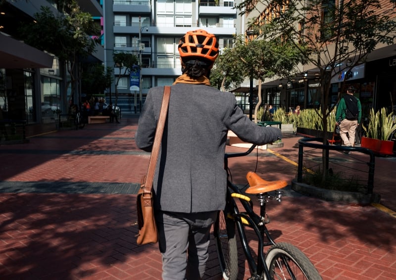 Person wearing an orange bike helmet and carrying a shoulder bag, walking a bicycle through a modern pedestrian walkway lined with trees and buildings.