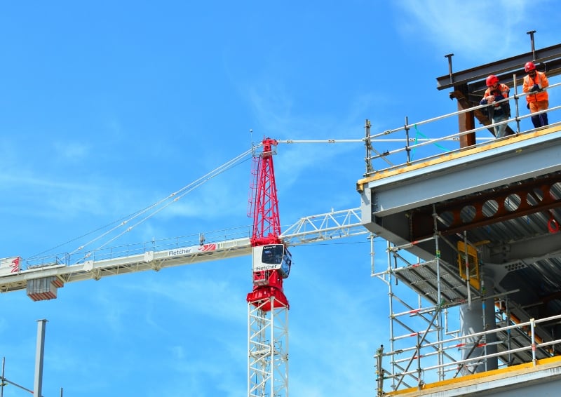 A photo of a construction crane against a blue sky in New Zealand. A building under construction is visible on the right, with a couple of workers standing on one of the floors, wearing personal protective equipment.
