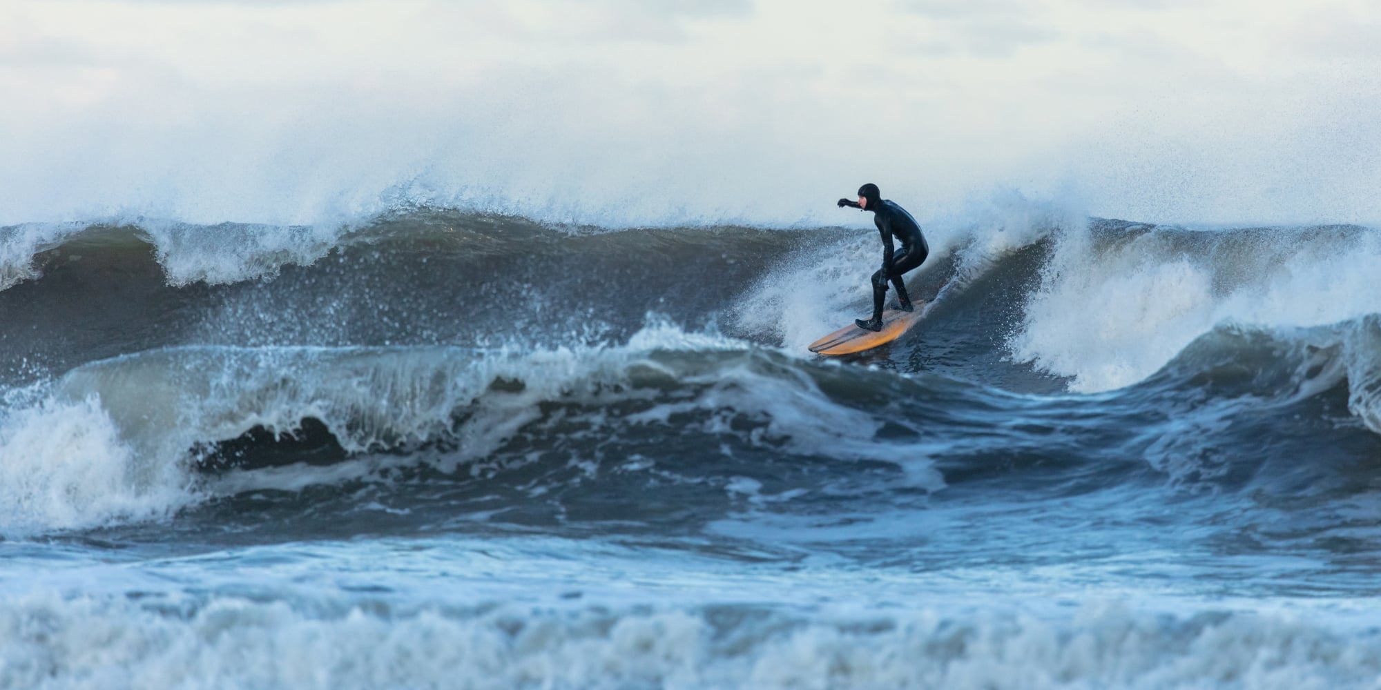 A photo of a person surfing a big wave in Tynemouth, UK. The sky is full of clouds.