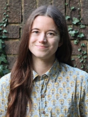 Head-and-shoulders portrait of a person with long hair wearing a patterned button-up shirt, standing in front of a brick wall with climbing ivy.