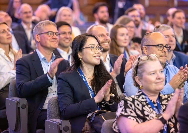 Audience seated in a conference auditorium applauding during a presentation, with attendees wearing business attire and event lanyards in a well-lit indoor venue.