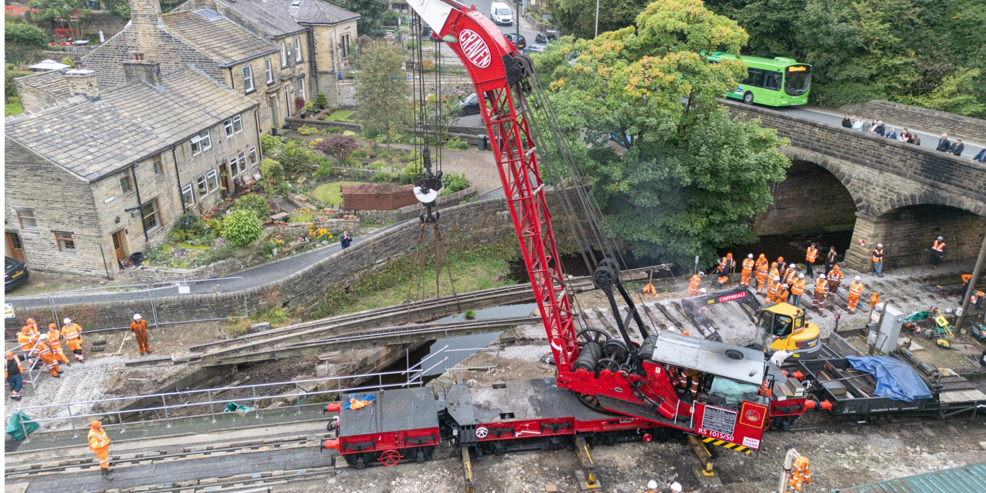 An aerial photo of a large red crane demolishing an old bridge over the Bridgehouse Beck tributary in west Yorkshire. The crane is the main feature of the image, but houses are visible on the left-hand side of the background, and a road with a bus on it on the right. There are a few people wearing personal protective equipment, including high-vis clothing, on the site.