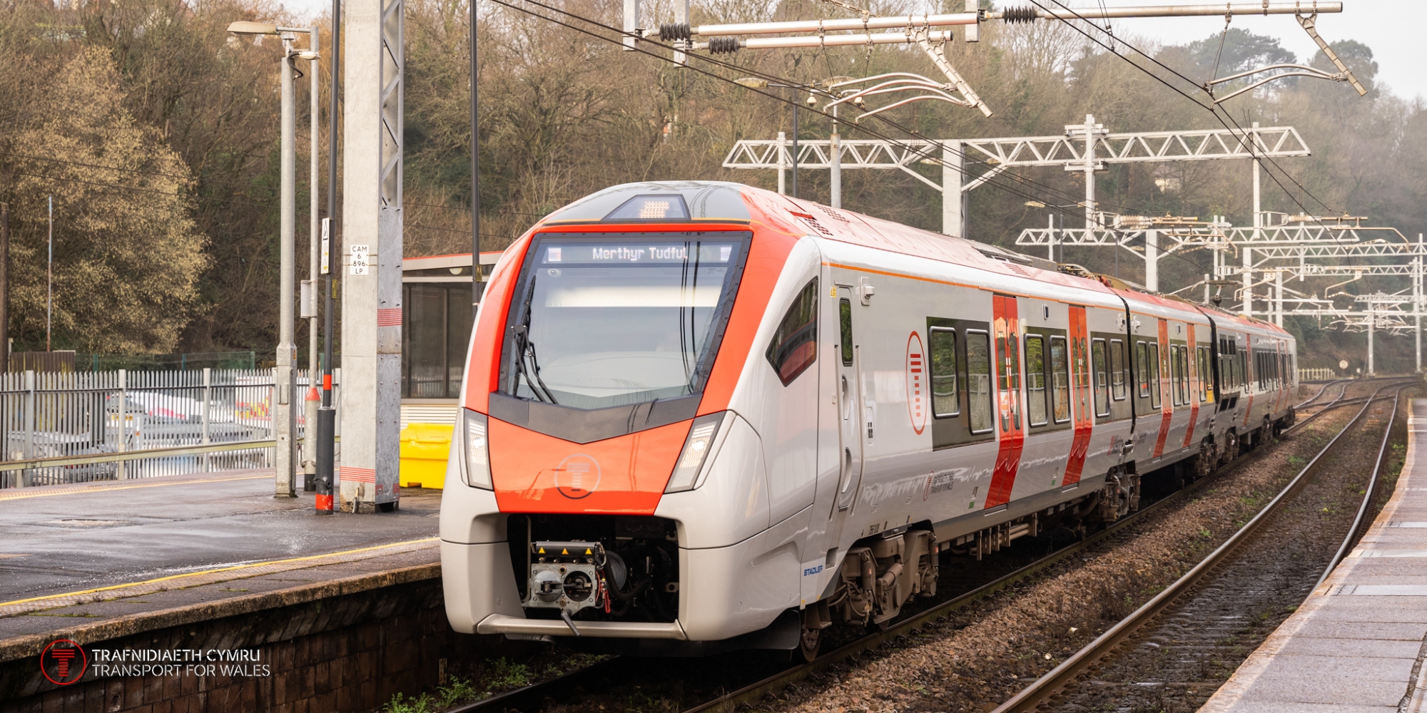 A Transport for Wales photo of a white train with orange accents waiting at an overground train station platform. The platform is empty. There are trees in the background and cables overhead.