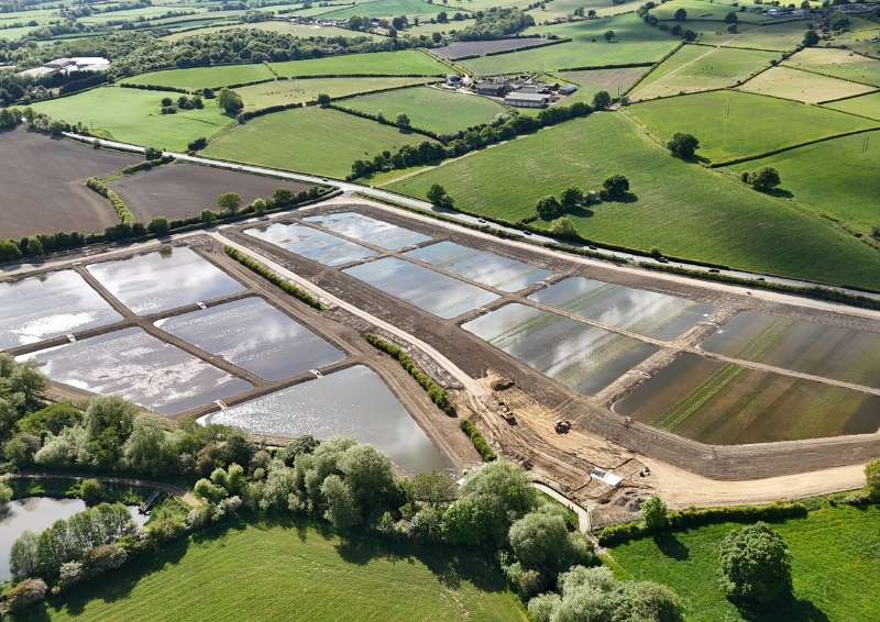 Aerial view of an integrated constructed wetland with rectangular water treatment ponds surrounded by green fields, trees, and rolling countryside