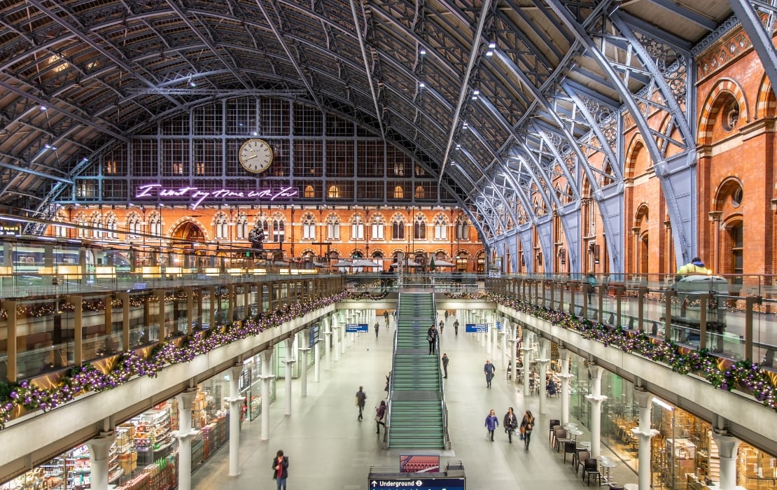 A photo of the interior of St Pancras International Station in London taken from the upper level, looking down at two rows of shops on either side, with a hall in the middle that passengers are walking through.