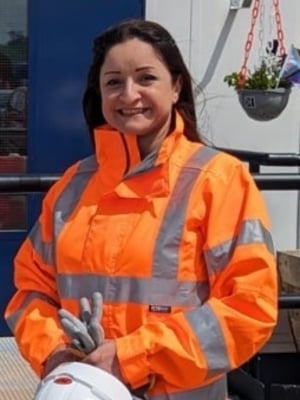 Portrait of a person wearing a bright orange high-visibility jacket and holding a white hard hat and protective gloves, standing outdoors in front of a site cabin.