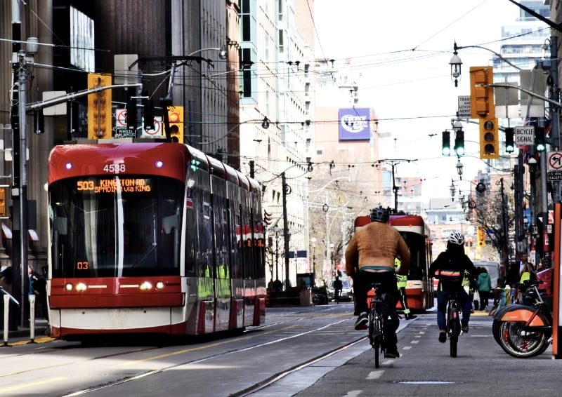 Red streetcar on King Street in downtown Toronto with cyclists riding in a dedicated bike lane, urban traffic lights and modern city buildings in the background.