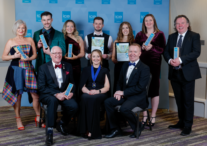 Group of nine people dressed in formal evening attire posing for a photo at an awards event. Several individuals are holding rectangular award trophies, and three are displaying framed certificates. The backdrop features a blue banner with the ICE logo repeated across it.