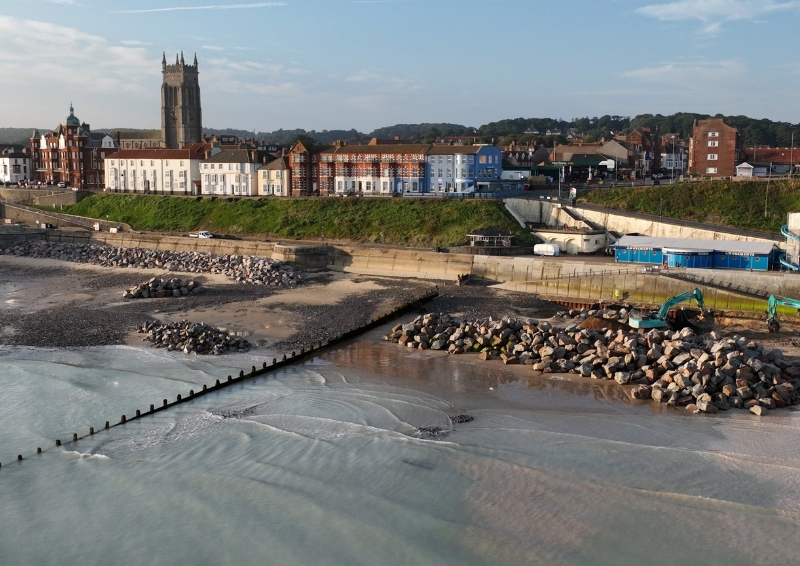 A birds-eye view photograph of rock revetments being built along Norfolk's coastline. The town is in the background, while rocks are being brought in by bright teal plant along the beach.