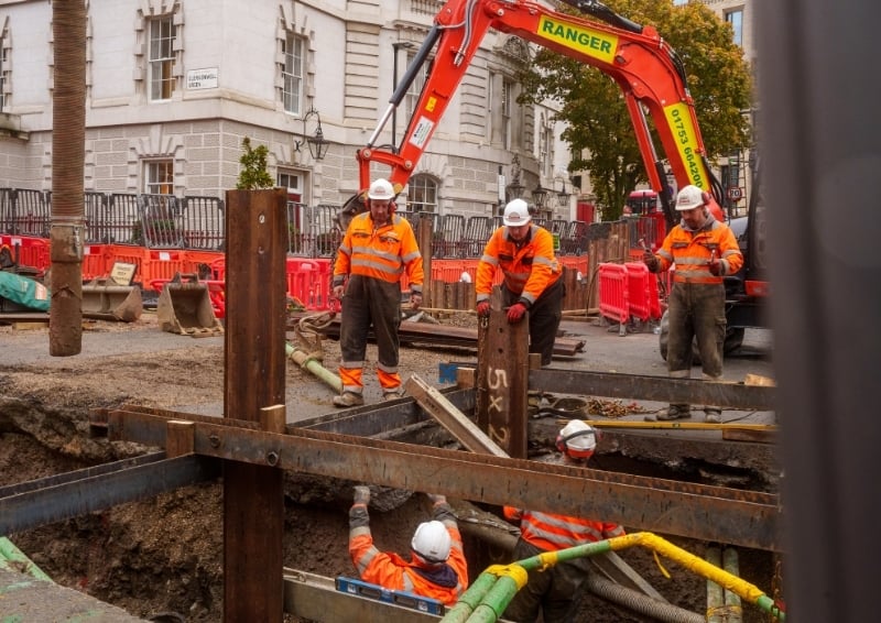 Construction workers in high-visibility orange safety gear operate at a street excavation site. They are handling large steel beams and equipment near an open trench, with one worker positioned inside the trench.
