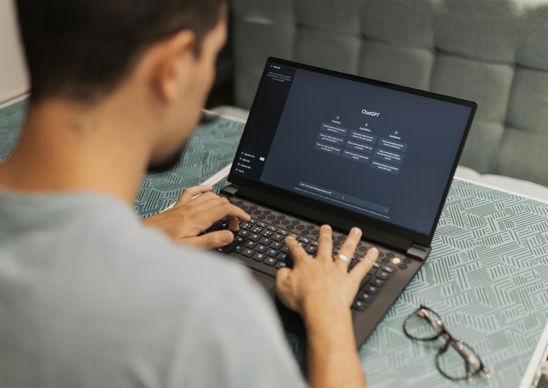 A person typing on a laptop at a table, with the screen displaying an AI chat interface. A pair of glasses rests on the table beside the laptop.