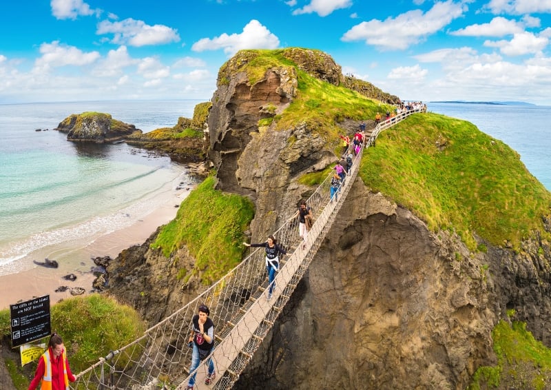 A scenic coastal view featuring the Carrick-a-Rede Rope Bridge in Northern Ireland. The rope bridge stretches between two rocky cliffs covered in green grass, with several people walking across it.