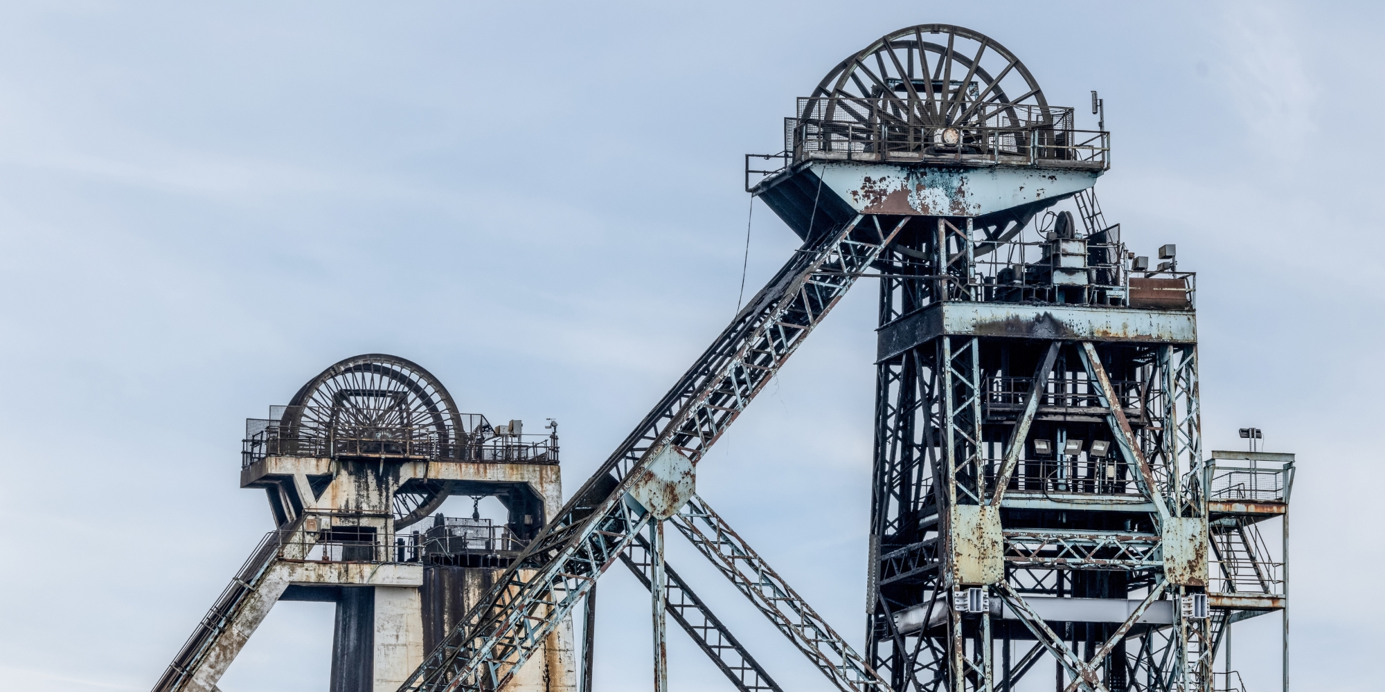 Two large, weathered steel mining headframes with rusted metal supports and machinery, connected by angled conveyor structures, set against a pale sky.